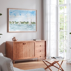 May include: A watercolor painting of sailboats on the water hangs above a wooden cabinet. The cabinet has a woven door and three drawers. A glass coffee table and white curtains are in the background.