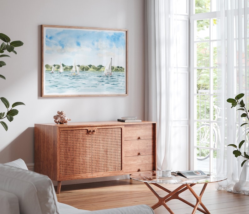 May include: A watercolor painting of sailboats on the water hangs above a wooden cabinet. The cabinet has a woven door and three drawers. A glass coffee table and white curtains are in the background.