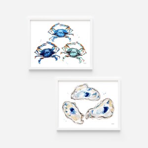 May include: Two framed watercolor paintings of blue crabs and oysters. The top painting features three blue crabs with orange claws. The bottom painting features four oysters with blue and white interiors.