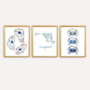 May include: A set of three framed wall art prints. The left print shows three watercolor oysters. The center print features a floral-patterned map of Maryland with the text Maryland below. The right print displays three blue crabs in a vertical row.