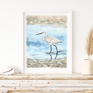 May include: Watercolor painting of a white egret standing in shallow water with its reflection visible. The egret has a long, thin beak and long legs. The water is a light blue color and the background is a light brown color.