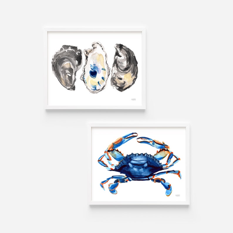 May include: Two framed watercolor paintings of coastal imagery. The top painting features three oyster shells in shades of gray, white, and blue. The bottom painting features a blue crab with orange claws.