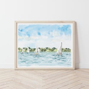 May include: Watercolor painting of sailboats on a lake under a blue sky with fluffy clouds. The artwork features several white sailboats with their sails up, sailing on blue water. Green trees line the horizon. The painting is framed in a light wood frame.