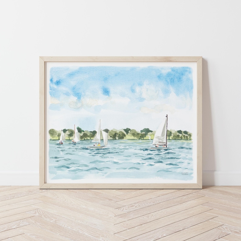 May include: Watercolor painting of sailboats on a lake under a blue sky with fluffy clouds. The artwork features several white sailboats with their sails up, sailing on blue water. Green trees line the horizon. The painting is framed in a light wood frame.