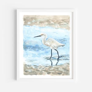 May include: Watercolor painting of a white egret standing in shallow blue water. The egret has a long, thin beak and long, slender legs. The water is clear and reflects the sky.