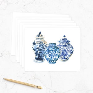 May include: A set of blank greeting cards featuring a watercolor illustration of three blue and white ginger jars. The jars have intricate floral and geometric designs. A gold pen rests on the white surface.