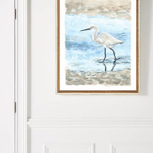 May include: A framed watercolor painting of a white egret standing in shallow water. The egret has a long, thin beak and long legs. The water is a light blue color and the background is a light brown color.