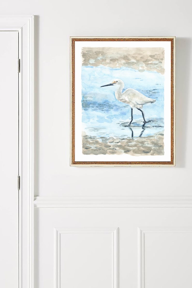 May include: A framed watercolor painting of a white egret standing in shallow water. The egret has a long, thin beak and long legs. The water is a light blue color and the background is a light brown color.