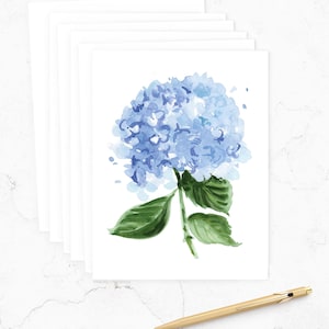 May include: A stack of white blank cards with a watercolor painting of a blue hydrangea flower and green leaves. A gold pen rests on the white surface. The floral art is a soft, delicate design.