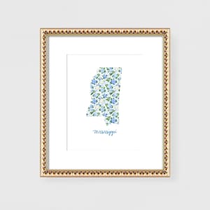May include: Framed art print featuring the state of Mississippi filled with a floral pattern of blue and white flowers. The word "Mississippi" is written in cursive below the state outline. The frame is gold with a decorative edge.