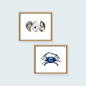 May include: Two framed watercolor paintings. The first painting features three oyster shells in shades of gray, white, and blue. The second painting features a blue crab with orange claws.