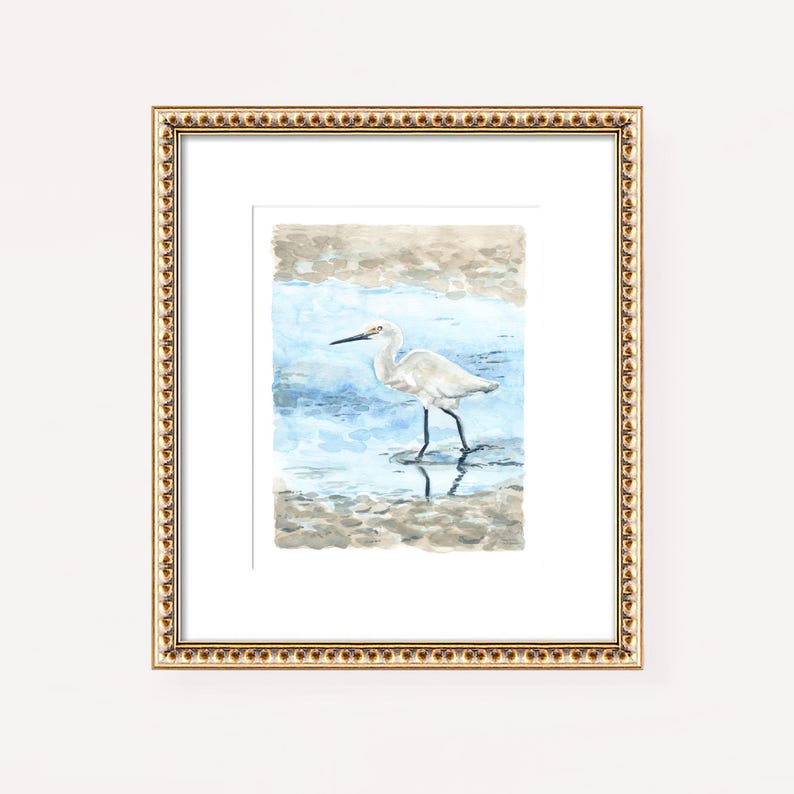 May include: A watercolor painting of a white egret standing in shallow water. The egret is facing left and has its head tilted down as if it is looking for food. The water is a light blue color and the background is a light brown color.