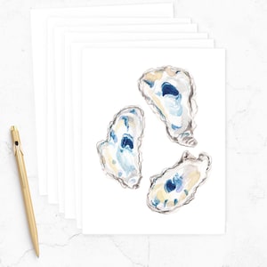May include: Watercolor art print featuring three oyster shells in shades of blue, white, and yellow. The artwork is on a white background, with a stack of blank white cards and a gold pen in the image.