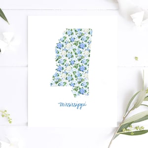 May include: A white print featuring the state of Mississippi filled with a blue and white floral pattern. The word "Mississippi" is written in cursive script in a light blue color at the bottom of the print.