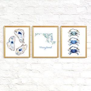 May include: Three framed wall art prints with gold frames. The left print shows three oyster shells. The center print displays a map of Maryland filled with blue and white floral patterns and the text Maryland. The right print features three blue crabs.