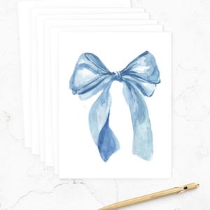 May include: A stack of white greeting cards with a watercolor illustration of a blue bow. A gold pen rests on the surface. The bow is painted in shades of blue, with delicate details and shading.