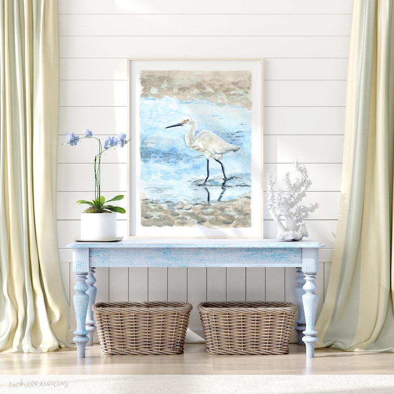 May include: A watercolor painting of a white egret standing in shallow water. The egret is facing left and has its wings folded. The water is a light blue color and the background is a light brown color.