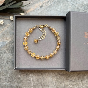 May include: A gold bracelet with a chain extender and a row of faceted, light brown gemstone beads. The bracelet is displayed in a gray box with a white label that says "Sara".