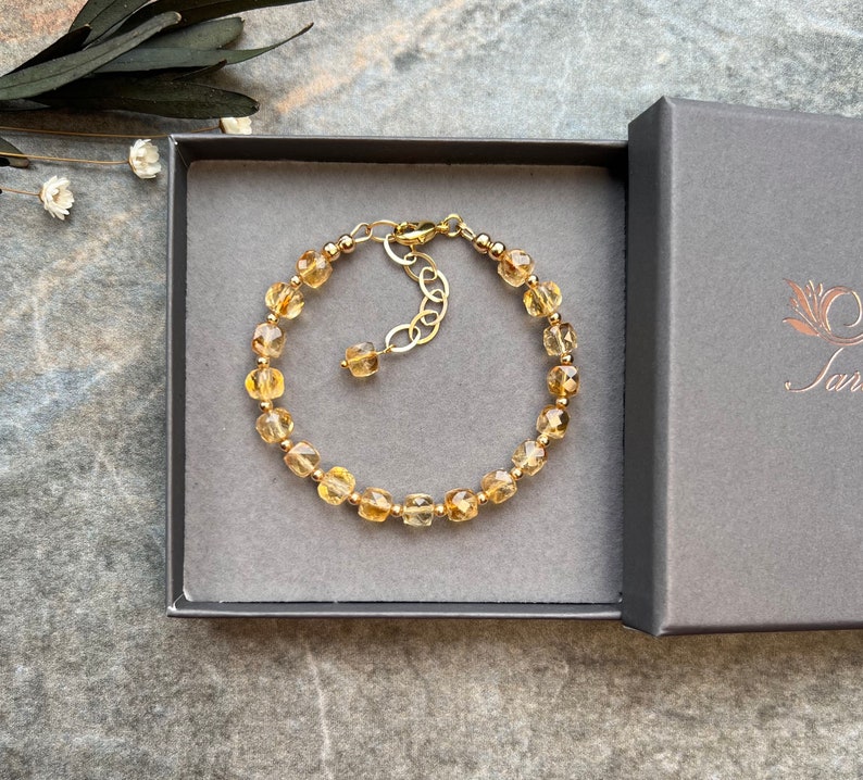May include: A gold bracelet with a chain extender and a row of faceted, light brown gemstone beads. The bracelet is displayed in a gray box with a white label that says "Sara".