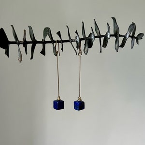 Puede incluir: Un par de pendientes de oro con colgantes de cubo de lapisl&aacute;zuli azul que cuelgan de una rama.