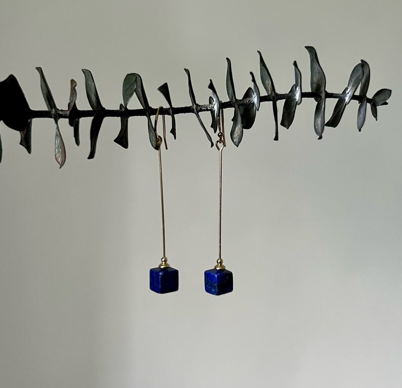 Puede incluir: Un par de pendientes de oro con colgantes de cubo de lapisl&aacute;zuli azul que cuelgan de una rama.