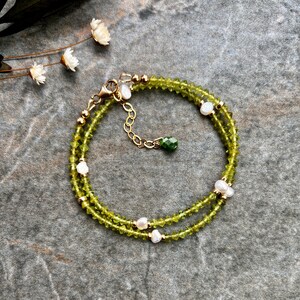 May include: A double-strand bracelet featuring faceted olive green beads and small white freshwater pearls. The design includes gold-toned metal beads, a lobster claw clasp, an adjustable chain, and a single faceted dark green bead charm.