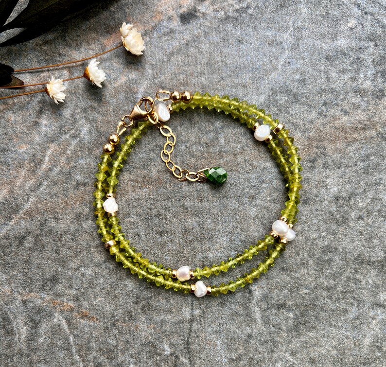 May include: A double-strand bracelet featuring faceted olive green beads and small white freshwater pearls. The design includes gold-toned metal beads, a lobster claw clasp, an adjustable chain, and a single faceted dark green bead charm.