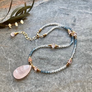 K&ouml;nnte beinhalten: Eine zarte Halskette mit einem gro&szlig;en, tropfenf&ouml;rmigen Rosenquarz-Anh&auml;nger. Die Halskette ist mit kleinen, klaren und blauen Perlen besetzt, die mit rosa und goldenen Akzenten durchsetzt sind. Der Verschluss und die Kette sind goldfarben und schaffen ein elegantes und feminines Accessoire.