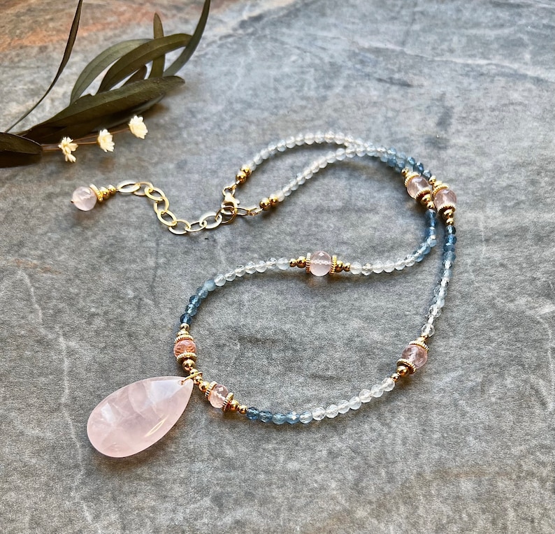 K&ouml;nnte beinhalten: Eine zarte Halskette mit einem gro&szlig;en, tropfenf&ouml;rmigen Rosenquarz-Anh&auml;nger. Die Halskette ist mit kleinen, klaren und blauen Perlen besetzt, die mit rosa und goldenen Akzenten durchsetzt sind. Der Verschluss und die Kette sind goldfarben und schaffen ein elegantes und feminines Accessoire.