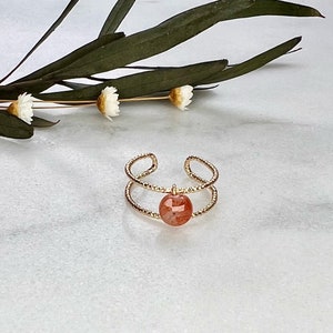Könnte beinhalten: Ein goldener Ring mit einem doppelten Band und einem runden, orangefarbenen Sonnensten-Anhänger.