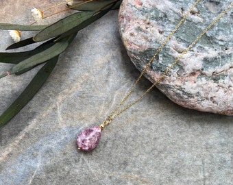May include: A delicate gold chain necklace featuring a single teardrop-shaped pendant made of a mottled, textured purple and white stone. The pendant is attached to the chain with a small gold-toned metal bead and a simple jump ring connector.