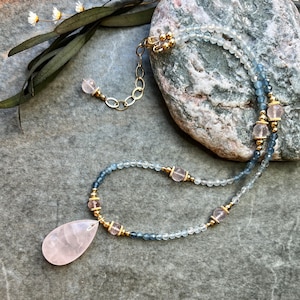 K&ouml;nnte beinhalten: Eine zarte Halskette mit einem tropfenf&ouml;rmigen Rosenquarz-Anh&auml;nger. Die Halskette enth&auml;lt kleine, hellblaue und klare Perlen, die mit goldenen Akzenten und rosa Edelsteinen durchsetzt sind. Der Verschluss und die Kette sind goldfarben.