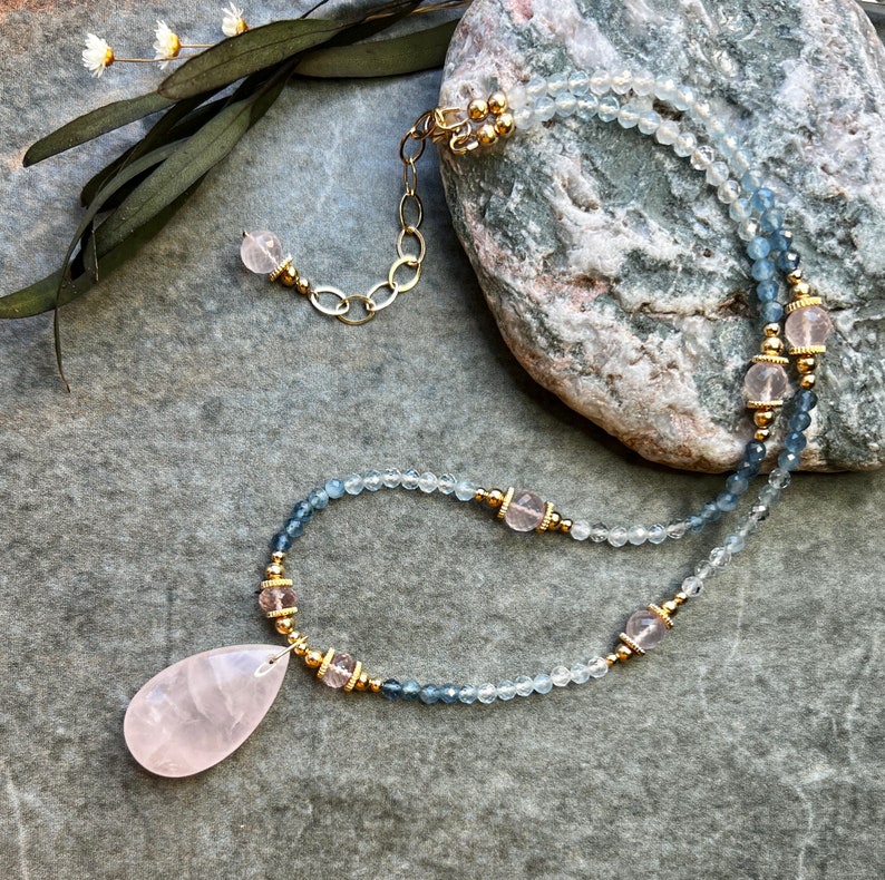 K&ouml;nnte beinhalten: Eine zarte Halskette mit einem tropfenf&ouml;rmigen Rosenquarz-Anh&auml;nger. Die Halskette enth&auml;lt kleine, hellblaue und klare Perlen, die mit goldenen Akzenten und rosa Edelsteinen durchsetzt sind. Der Verschluss und die Kette sind goldfarben.