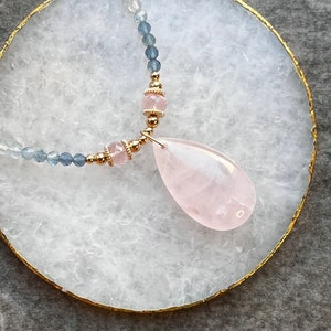 K&ouml;nnte beinhalten: Eine zarte Halskette mit einem tropfenf&ouml;rmigen Rosenquarz-Anh&auml;nger. Die Halskette hat eine goldene Kette mit kleinen blauen und klaren Perlen und rosa Edelsteinakzenten. Der Anh&auml;nger hat eine zarte rosa Farbe.