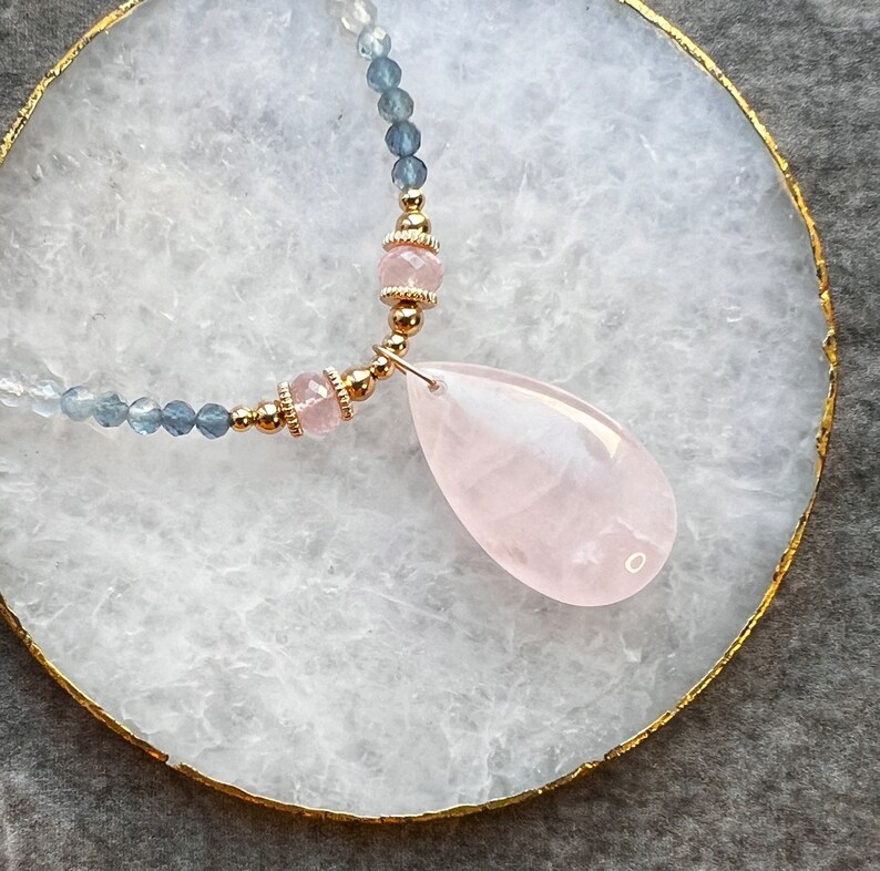 K&ouml;nnte beinhalten: Eine zarte Halskette mit einem tropfenf&ouml;rmigen Rosenquarz-Anh&auml;nger. Die Halskette hat eine goldene Kette mit kleinen blauen und klaren Perlen und rosa Edelsteinakzenten. Der Anh&auml;nger hat eine zarte rosa Farbe.
