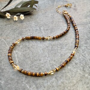 Könnte beinhalten: Eine zarte Halskette mit einer Kombination aus kleinen, rechteckigen Tigeraugenperlen und goldenen Akzenten. Die Halskette enthält auch ovale, durchscheinende gelbe Perlen. Die Kette ist goldfarben und die Halskette wird auf einer grauen Oberfläche präsentiert.