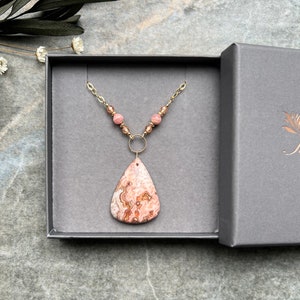 May include: A gold chain necklace with a pink and brown agate pendant and pink beads. The necklace is displayed in a grey jewellery box.