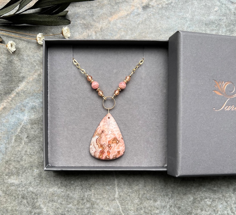 May include: A gold chain necklace with a pink and brown agate pendant and pink beads. The necklace is displayed in a grey jewellery box.