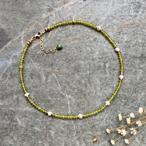 May include: A necklace featuring a strand of small, faceted olive-green beads interspersed with irregular white freshwater pearls. The piece includes a gold-toned metal clasp, an adjustable chain, and a small, teardrop-shaped green stone charm.