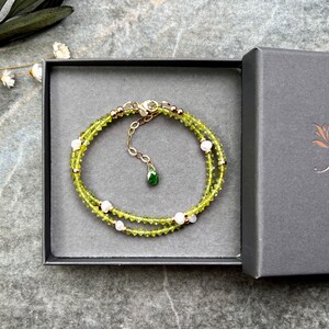 May include: A delicate bracelet featuring small, faceted lime-green peridot beads interspersed with tiny white pearls and gold-toned metal spacers. The bracelet includes a gold-toned chain extender with a small, teardrop-shaped green gemstone charm.
