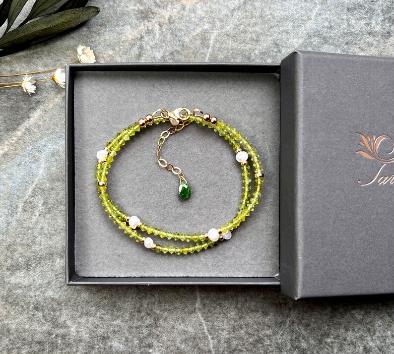 May include: A delicate bracelet featuring small, faceted lime-green peridot beads interspersed with tiny white pearls and gold-toned metal spacers. The bracelet includes a gold-toned chain extender with a small, teardrop-shaped green gemstone charm.
