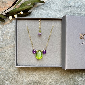 May include: A gold necklace with a green pear-shaped gemstone pendant and three smaller purple gemstones. The necklace is in a grey box with a white label that says "Jari".