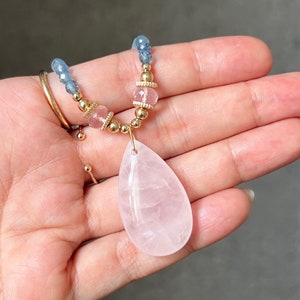 K&ouml;nnte beinhalten: Eine zarte Halskette mit einem gro&szlig;en, tropfenf&ouml;rmigen Rosenquarz-Anh&auml;nger. Die Halskette enth&auml;lt kleine, runde goldene Perlen, rosa und blaue Edelsteinperlen und goldene Akzente. Der Schmuck wird in einer Hand gehalten.
