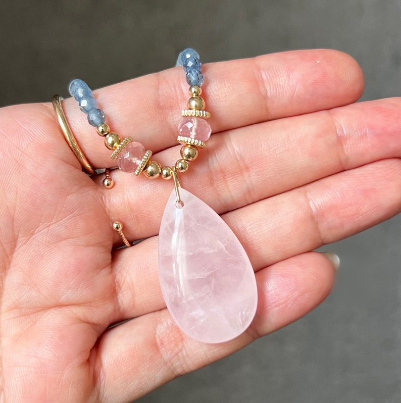 K&ouml;nnte beinhalten: Eine zarte Halskette mit einem gro&szlig;en, tropfenf&ouml;rmigen Rosenquarz-Anh&auml;nger. Die Halskette enth&auml;lt kleine, runde goldene Perlen, rosa und blaue Edelsteinperlen und goldene Akzente. Der Schmuck wird in einer Hand gehalten.