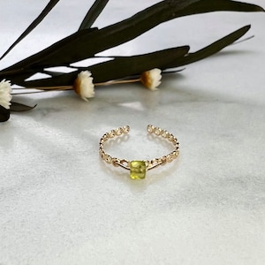 May include: A delicate gold ring with a small, square, light green gemstone in the center. The ring is made of a chain-like design.