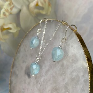 May include: A silver necklace and earrings set featuring light blue aquamarine heart-shaped stones. The necklace has a delicate chain and a pendant with a heart-shaped aquamarine stone. The earrings are also heart-shaped and have a simple design.