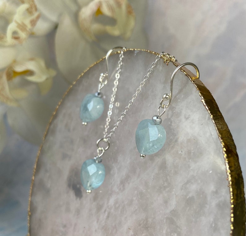 May include: A silver necklace and earrings set featuring light blue aquamarine heart-shaped stones. The necklace has a delicate chain and a pendant with a heart-shaped aquamarine stone. The earrings are also heart-shaped and have a simple design.