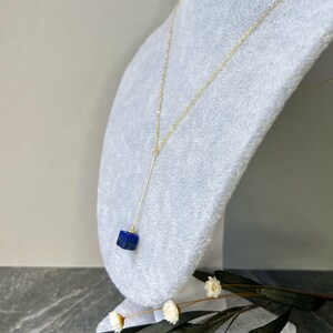 Puede incluir: Un collar de cadena dorada con un largo y delgado colgante dorado y una piedra preciosa c&uacute;bica azul oscuro.