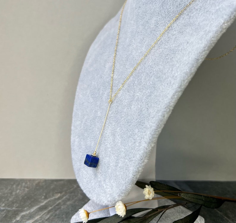 Puede incluir: Un collar de cadena dorada con un largo y delgado colgante dorado y una piedra preciosa c&uacute;bica azul oscuro.