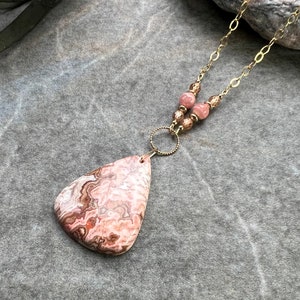 May include: A pink and brown agate pendant necklace with a gold chain and brown beads. The pendant is a teardrop shape with a swirling pattern.